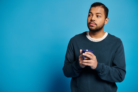 thoughtful african american man in 20s holding cup in hands and looking to side on blue background
