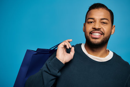 portrait of cheerful african american young man with shopping bag over shoulder on blue backgroundの写真素材
