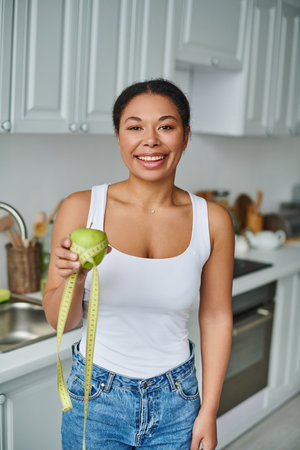 happy african american woman with measuring tape and apple promoting a healthy diet in kitchenの写真素材