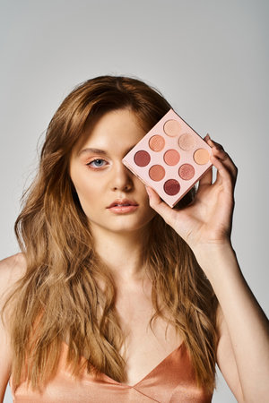 Beauty shot of woman holding peach makeup palette near eye on grey studio backgroundの写真素材