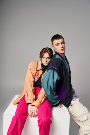 appealing woman in fashionable bomber looking at camera next to her handsome boyfriend on white cube