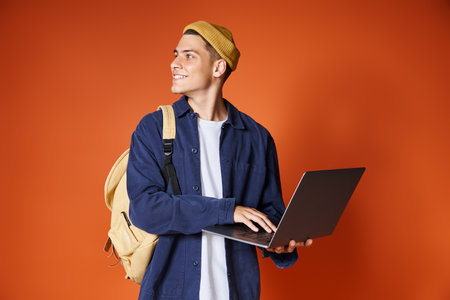 smiling student with backpack networking to laptop and looking to side against terracotta background