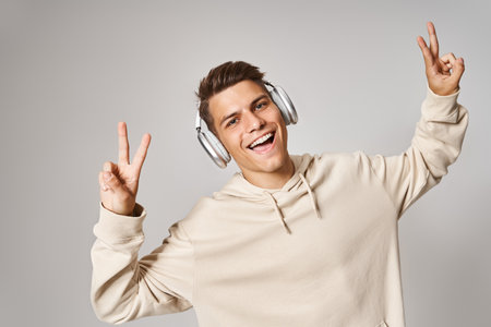 cheerful student in headphones smiling and showing sign peace with hands against light background