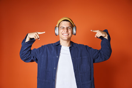 cheerful young man in yellow hat pointing with fingers to headphones against terracotta background