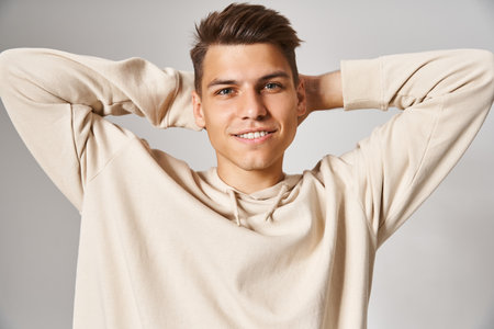 charismatic man in white hoodie smiling and putting hands behind head against light background