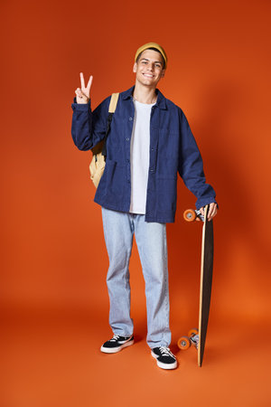 attractive young man standing with skateboard and showing peace on terracotta backgroundの写真素材