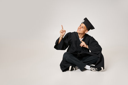 cheerful young man in graduate gown and cap sitting and showing with fingers to upの写真素材