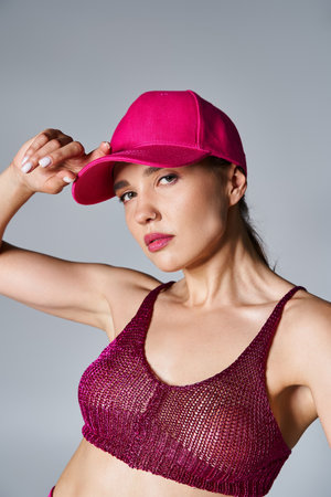 Portrait of fancy brunette woman in red top and touching pink cap with one hand on grey backdropの写真素材