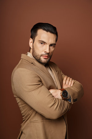 handsome man with bristle posing in elegant attire standing with folded arms on beige backgroundの写真素材