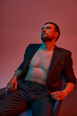 handsome man with bare chest posing in pinstripe suit and sitting in studio with red and blue lightの写真素材
