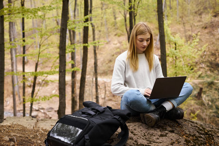Young blonde woman with backpack hiking in woods, sitting on boulder with laptop and workingの写真素材
