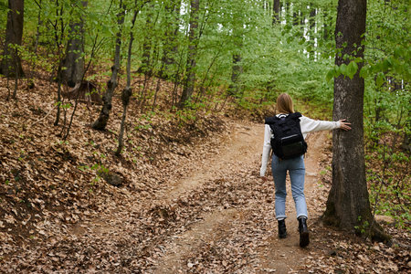 Back full length view of blonde woman hiking walking in green forest touching trees