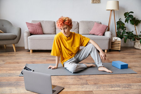 beautiful queer person in yellow t shirt sitting on yoga mat and looking at laptop before exercisingの写真素材