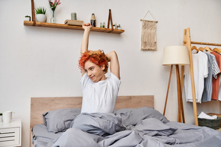 extravagant young queer person in casual attire waking up and stretching in her bed, leisure timeの写真素材