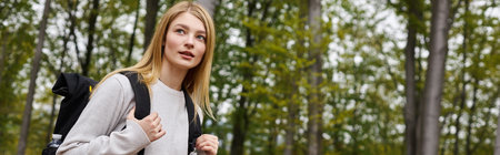 banner of curious blonde woman holding backpack, wearing sweater, looking through the woods