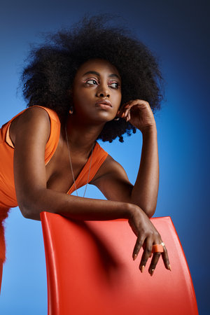 dreamy african american woman in peach fuzz dress posing next to chair on vivid blue backgroundの写真素材