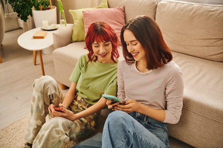 happy lesbian couple smiling and using smartphones near sofa, modern lifestyle and social media