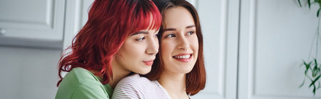 portrait of joyful and pierced lesbian woman with red hair embracing her girlfriend at home, bannerの写真素材