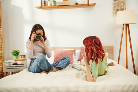 Candid home photo session of lesbian woman taking photo on retro camera of her girlfriend on bedの写真素材