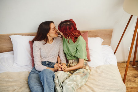 pleased young lesbian couple in casual wear holding hands and going to kiss while sitting on bedの写真素材