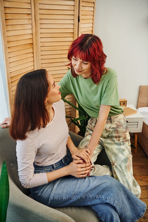 young and happy lesbian woman with red hair looking at her girlfriend sitting in armchair at homeの写真素材