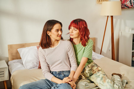 Young lesbian couple in casual attire sharing cozy moment and looking at each other in sunny bedroomの写真素材