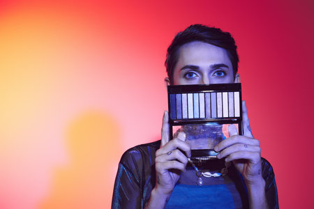 enticing gay man in casual attire using eyeshadows palette and looking at camera on vibrant backdrop