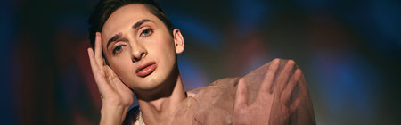 chic androgynous man with dark hair in pastel attire looking at camera on vivid backdrop, banner