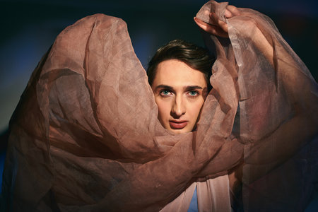 voguish androgynous fashionista with dark hair in pastel attire looking at camera on vivid backdropの写真素材