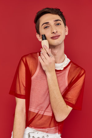 cheerful appealing androgynous man with red fishnet holding makeup foundation and looking at cameraの写真素材
