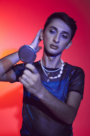 alluring androgynous man in vivid attire looking at small mirror while on red and orange backdropの写真素材