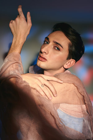 elegant androgynous fashionista with dark hair in pastel attire looking at camera on vivid backdropの写真素材