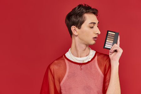 fashionable androgynous man with stylish red fishnet holding eye shadows palette and looking awayの写真素材