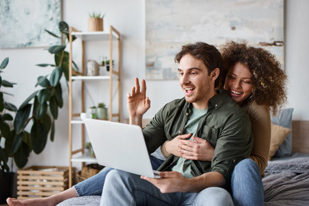 In their cozy bedroom, curly young woman and brunette man talking online on their laptopの写真素材
