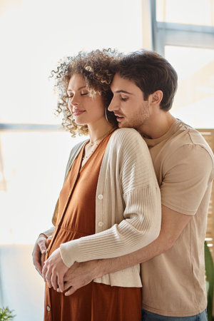 Expecting baby couple, man hugging his pregnant wife from behind standing near windowの写真素材