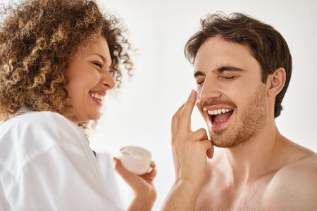 Woman applying cream to her man in bathroom and laughing together, cheerful happy coupleの写真素材