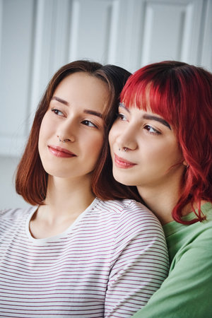 portrait of joyful and pierced lesbian couple looking away and spending time together at homeの写真素材
