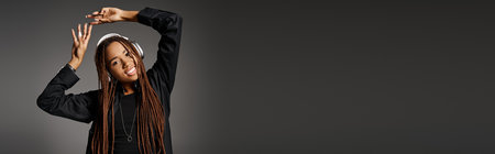 happy and young African American woman with headphones dancing to music on grey backdrop, banner