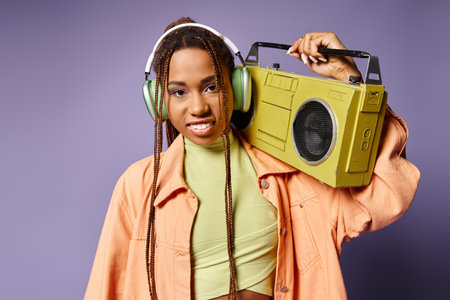 happy african american woman in wireless headphones standing with retro boombox on purple backdrop