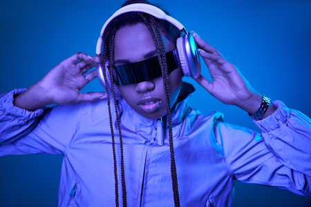pretty dark skinned woman posing in stylish sunglasses with headphones on blue with neon lightの写真素材