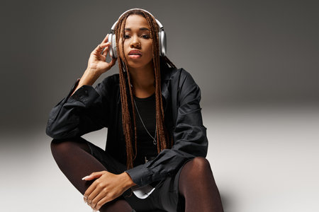 young african american woman in 20s with dreadlocks enjoying music in stylish headphones on greyの写真素材