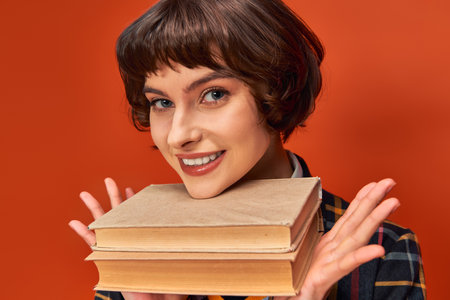 portrait of smiling college girl in uniform holding books near chin on orange background, knowledgeの写真素材