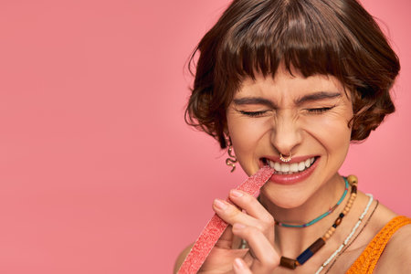 expressive woman with nose and white teeth biting sweet and sour candy strip on pink