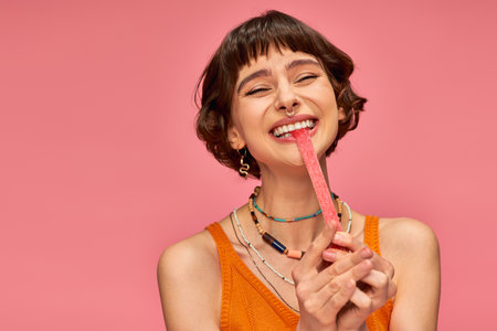 excited and young woman with nose piercing biting sweet and sour candy strip on pink background