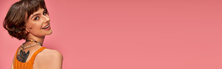 happy tattooed woman in 20s with nose piercing and brunette short hair smiling on pink, banner