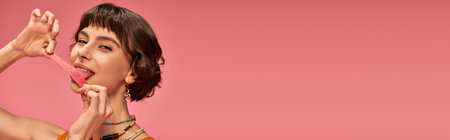 happy girl with nose piercing licking sweet and sour candy strip on pink background, horizontalの写真素材