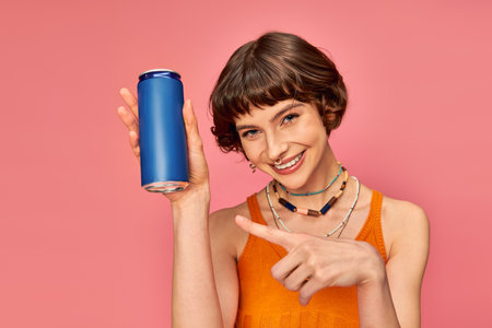 joyful young woman with short brunette hair pointing at soda can on pink, summer beverageの写真素材