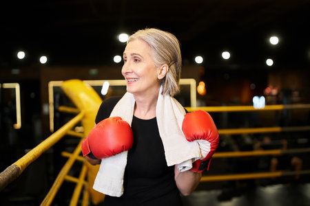 athletic gray haired senior woman with towel and boxing gloves looking away while on ring in gymの写真素材