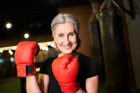 cheerful pretty senior sportswoman with gray hair in boxing gloves smiling at camera while in gymの写真素材