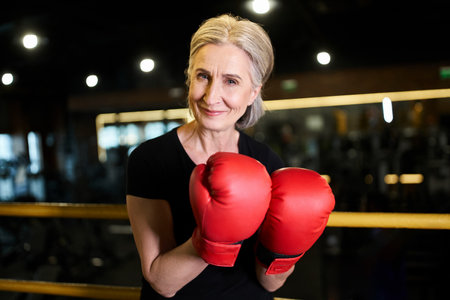 attractive joyful senior sportswoman with boxing gloves smiling at camera while on ring in gym